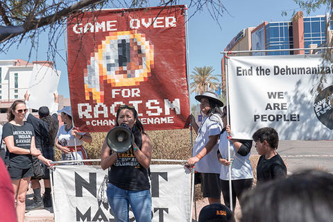 Imagining the Indian. Woman on stage wearing black tee and blue jeans holding a bullhorn in front of a huge banner reading: Game Over for Racism. 