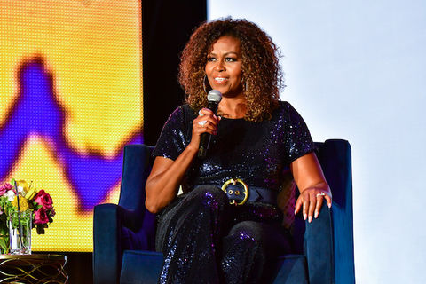 Michelle Obama sits on a blue chair on stage holding a microphone near her face.