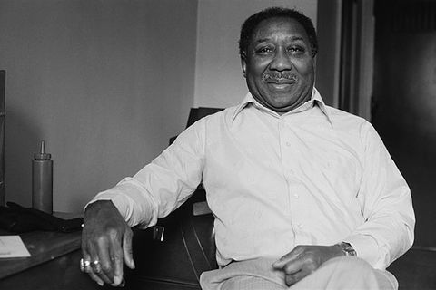 Muddy Waters. Black and white archival photo of Black man with short dark hair wearing white shirt and tan pants.