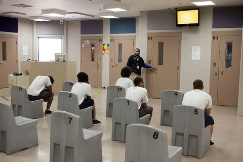 A group of young Black boys wear white t-shirts and sit in chairs with their backs to the camera. 