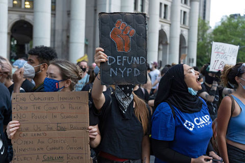 A protester with a scarf covering his face is seen displaying placard reading "Defund NYPD" 