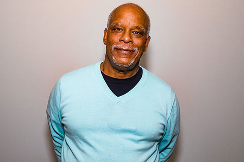 Stanley Nelson. Middle aged Black man with bald head and gray beard wearing a teal colored sweater and black Tee.