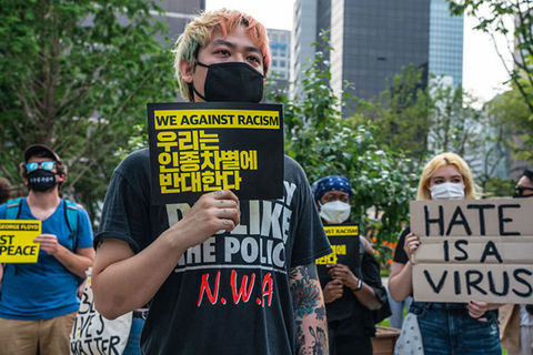 Stop AAPI Hate. Asian man with colorful hair wearing black mask and NWA Tee holding a sign reading "We against racism."