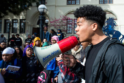 A Black student is holding a megaphone leading a crowd of students in protest.