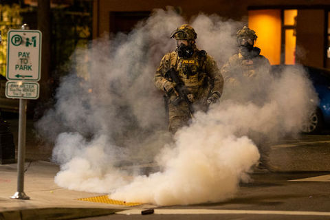 A federal officer wearing military gear walks in a city street surrounded by tear gas.