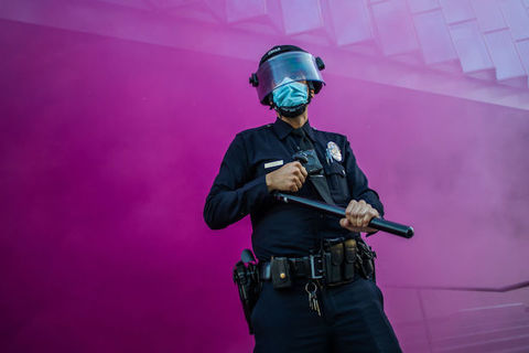 A uniformed police officer stands in front of a purple wall of smoke as they hold a baton and cover their face with a mask and shield. 