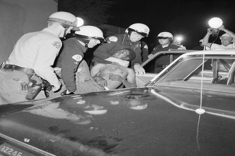Black and white photograph of police officers forcing a Black man into a police car during second night of the Watts rebellion.