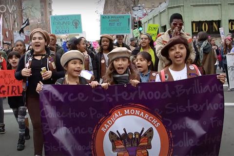 We Are the Radical Monarchs. Three young girls of color wearing scouts caps and carrying a purple banner reading "Radical Monarch" march in a rally.