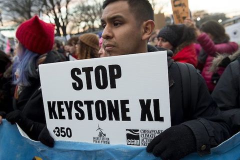 A shot of a young brown man standing outside at a protest and holding a sign that reads "Stop Keystone XL."