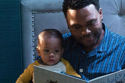 Black-ish. Black man with short dark hair reading children's book to Black baby boy.