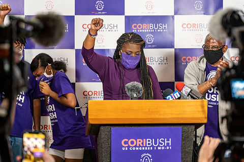 Cori Bush. Black woman wearing purple mask and top with a raised fist a podium surrounded by cameras.