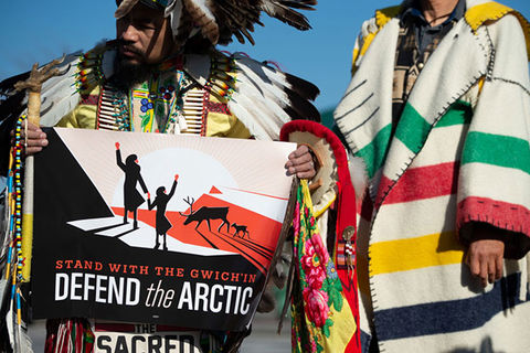 Defend the Arctic Protest. Indigenous man standing next to someone in colorful stripped coat, holds sign reading: Defend the Arctic.