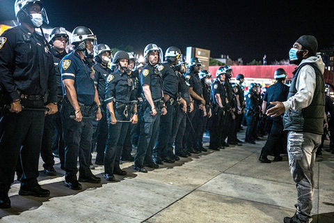 A Black protester wearing a mask stands in front of a blockade of police officers in riot gear.