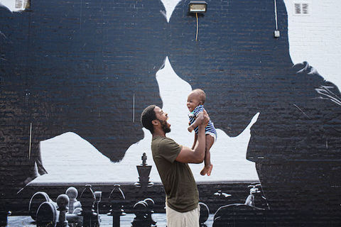 A Black man is holding a baby up in the air in front of a painted mural with a father and son.