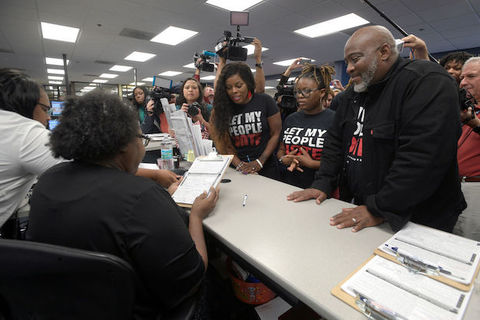 One Black man and two black women stand in front of a desk waiting on paperwork while wearing t-shirts that read, Let My People Vote. 