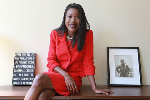 Isabel Wilkerson. Black woman with long dark straight hair wearing red skirt suit sitting between two small pieces of desk art,