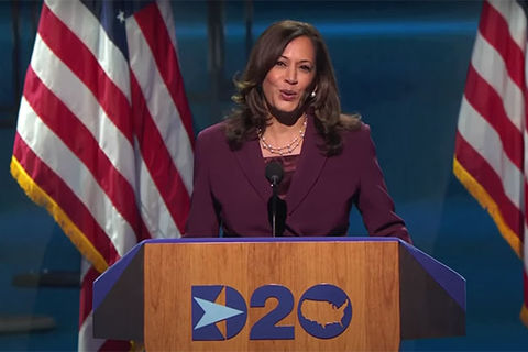 Kamala Harris. Black woman with shoulder length brown hair wearing maroon-colored suit standing in front of U.S. flags on stage.