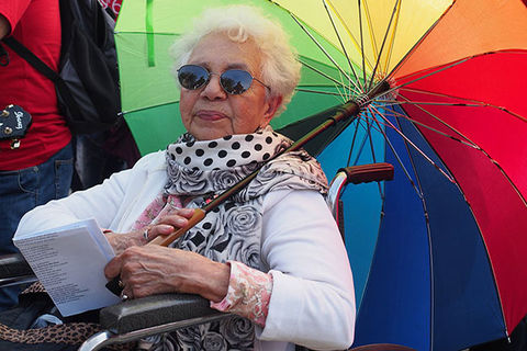 LGBTQ ELDER. Brown elder woman in a wheelchair with short gray hair holding a rainbow colored umbrella.