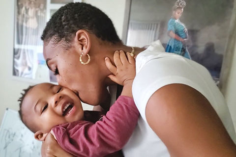 Lyft. Black woman with short dark hair wearing white Tee holding Black baby boy wearing maroon shirt.