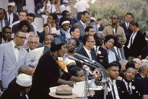 Mahalia Jackson. Black woman on stage in front of microphone surrounded by men in suits, including Martin Luther King, Jr.
