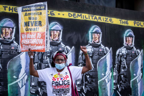 A  protester wearing a mask holds a raised black power fist holds a sign that reads, "American was never great! We need to Overthrow this system!" in front of a mural with NYPD officers in full riot gear and the banner above them that says, "Demilitarize The Police" 
