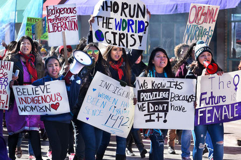 A group of Indigenous women march and hold colorful signs calling for justice. 