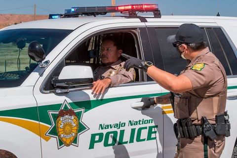 A uniformed officer stands and talks to another officer who sits inside a car that reads, Navajo Nation Police