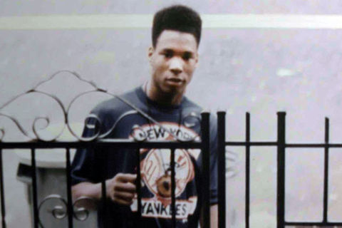 Yusuf Hawkins. Black teenage boy with high top wearing dark tee standing in front of a gate.