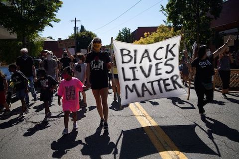 A woman wearing a black short holds hands with a young girl wearing a pink shirt. They are marching with a crowd at a peaceful protest as they hold a sign that reads, Black Lives Matter.