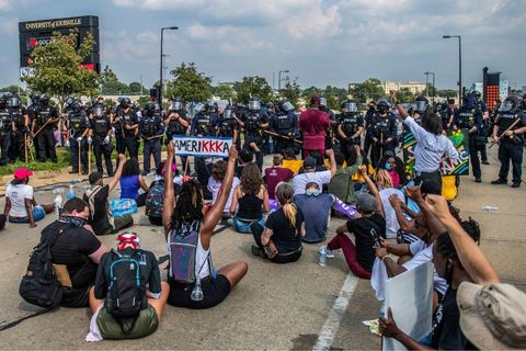 A row of police in riot gear -- batons and shields -- stand over a about 30 protesters sitting on the ground