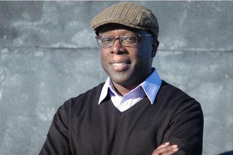 Middle age Black man smiling with paperboy cap, black sweater, and dress shirt underneath