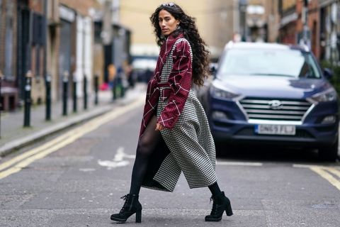 Brown, ambiguous woman walks across a cobblestone street with a long gray coat with cranberry sleeves