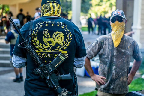 white man with his back to the camera wears a jacket that reads Columbia River Proud Boys. Another white man facing the camera has a yellow bandana covering his mouth and nose. 