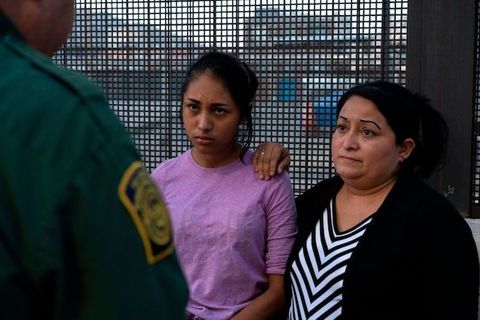 a young girl wearing a purple shirt stands next to her mother. They both look somber as they face a customs and border patrol agent, who stand with his back to the camera. 