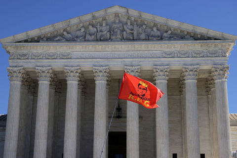 The Supreme Court stands in the background as a red flag with a drawing of Justice Ruth Bader Ginsberg and the words Notorious RBG flies in the foreground.