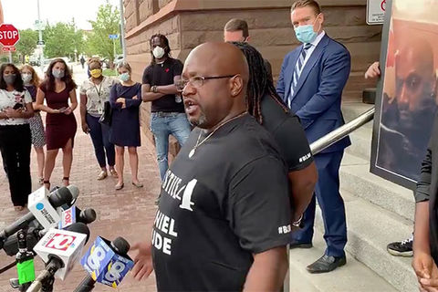 Joseph-Prude. Black man with bald head, glasses wearing black Tee stands in front of reporter's mics.