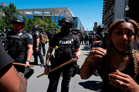 Kentucky-Protests. Black woman with long braids wearing black tank and black face mask moves ahead of a row of police officers holding batons.