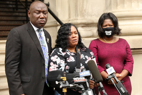 A Black man and two Black women, one wearing a mask that says Breonna Taylor, stand in front of a microphone. 