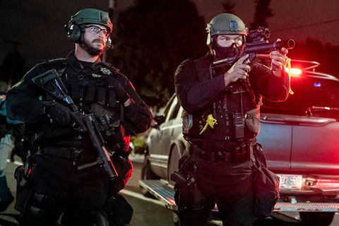 Police Violence. Two officers in uniform wearing helmets and carrying weapons while one is aiming the weapon.