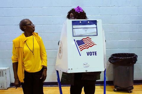 A Black woman wearing a yellow shirt and blazer speaks to another Black woman who wears a purple flower in her hair and stands behind a voting booth. 