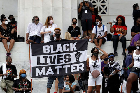 A large group of people are seated on a platform behind a Black man who stands and holds a large Black Lives Matter sign.