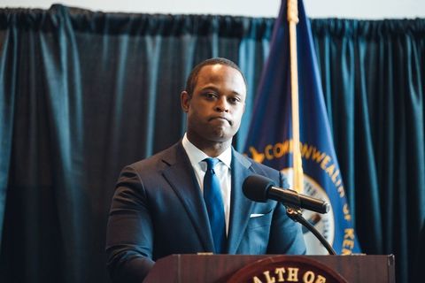 Black man with brown skin and close cropped hair in a blue suit standing at a lectern with a somber expression.