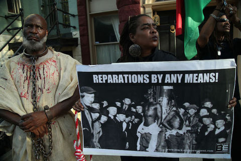 Three Black people hold a large protest sign that reads "Reparations By Any Means!"