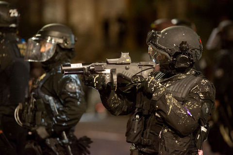 Police officers in black riot gear stand in the middle of a street as one points a gun in front of them.