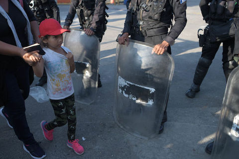 A small girl wearing a red hat hold hands with an adult as they walk past officers wearing riot gear. 
