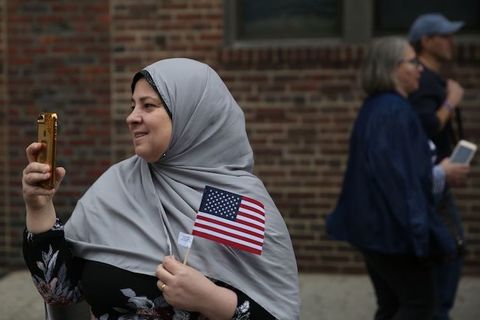 A woman wears a gray hijab smiles as she holds a small American flag and a smartphone.