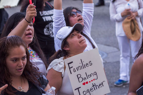 A woman wearing a white baseball cap looks to the sky and holds a sign that reads, Families Belong Together. 