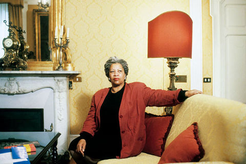 Toni Morrison. Black woman with long gray locs seated on a yellow sofa, wearing a black dress and red jacket. 