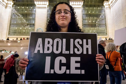A woman with dark curly hair holds a large sign that says, Abolish ICE