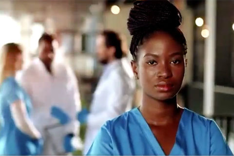 Black Coalition of docs. Young Black woman with braids in top bun wearing blue medical scrubs.